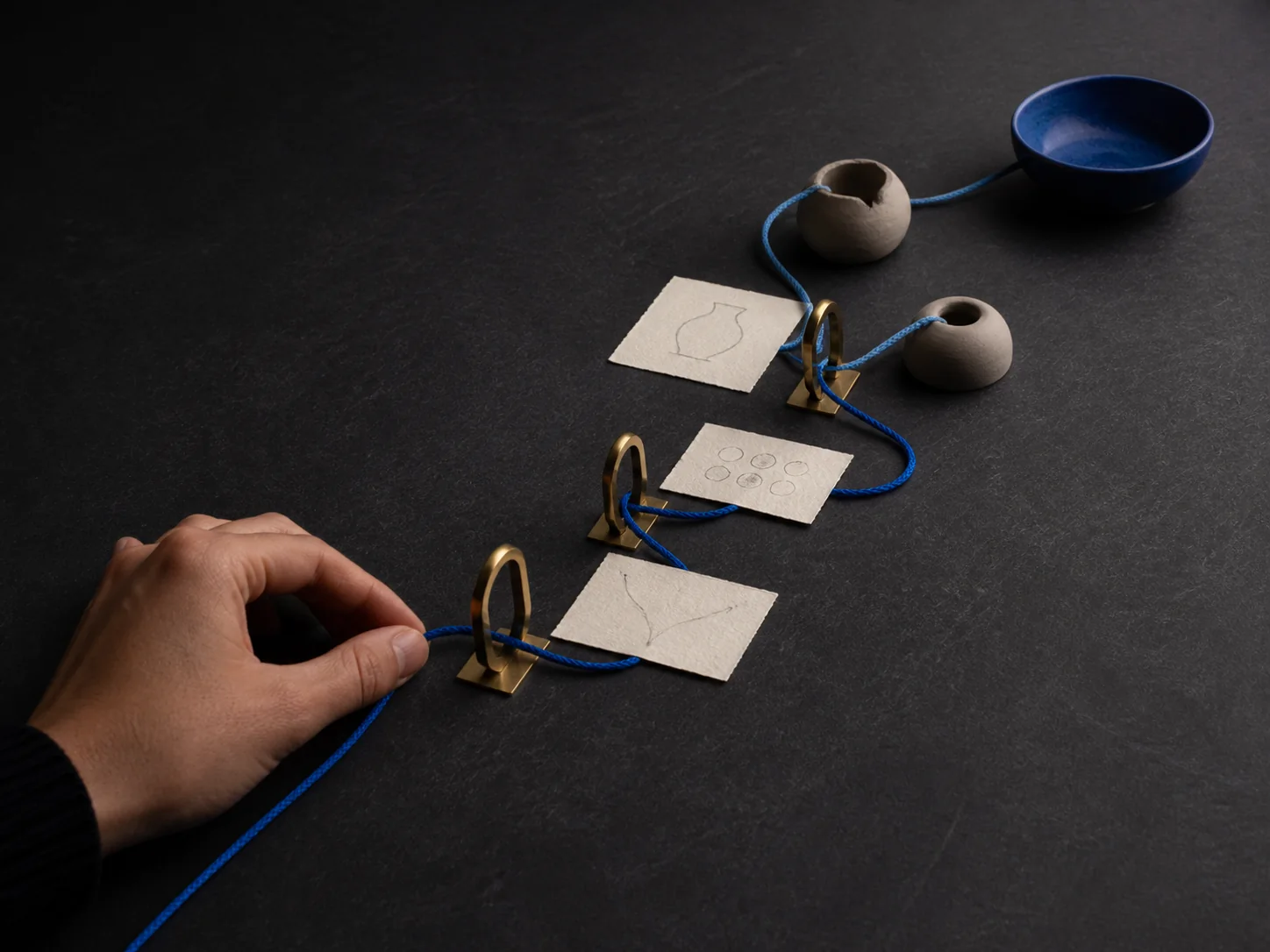 A hand guiding a blue cord through a series of brass gates toward finished forms